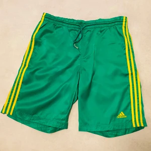 Retro Gröna shorts från Adidas - Snygga ovanliga gröna shorts från Adidas med gula ränder längs sidorna. Dragsko för justerbar passform. Skönt glansigt material! Storlek M