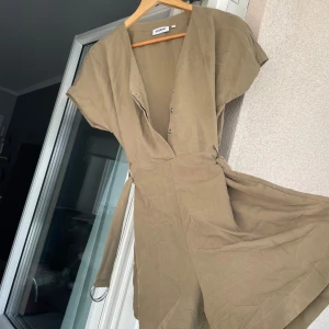 Beige/mossgrön klänning från Weekday - Säljer en stilren beige/mossgrön klänning från Weekday. Klänningen har en v-ringad design med korta ärmar och knappar framtill. Använt fåtal gånger och därför i mycket bra skick.