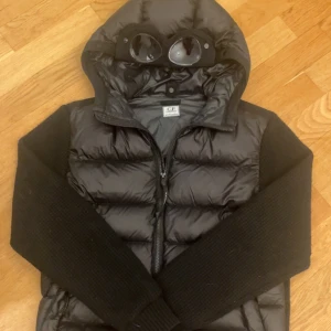 Svart Goggle Jacket från C.P. Company - Säljer en svart Goggle Jacket från C.P. Company. Jackan har en unik design med inbyggda glasögon i huvan och dragkedja framtill. Den är långärmad med quiltade detaljer och är tillverkad i syntetmaterial. Perfekt för dig som vill ha en stilren och funktionell jacka.