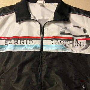Tracksuit jacak från Sergio Tacchini - Säljer en fräsch tracksuit jacka från Sergio Tacchini med dragkedja och färgglada ränder i rött och blått. Jackan har ett stort tryck av märkesloggan på bröstet och en klassisk krage. Köpt i Nice. Storleken är 164 tonåring 15-16 år.