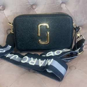 Svart axelväska från Marc Jacobs - Snygg svart axelväska från Marc Jacobs med guldiga detaljer och en bred axelrem med logotyp. Väskan har en dragkedja upptill och är perfekt för att bära dina nödvändigheter med stil.