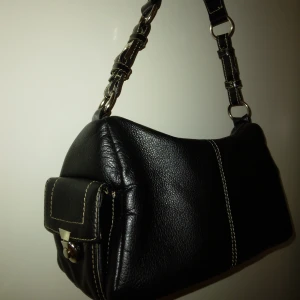 Svart snygg präglad handväska med flera fack. Fint skick. David Jones. - Svart handväska i präglat immitationsläder med silverfärgade detaljer och handtag/axelrem.  Från David Jones. Väskan har bla 2 små fack med stängbara lock på sidorna, samt extrafack på utsidan av väskan, I bak, samt några fack inuti väskan. Tygfodrad insida.  Fint skick. Mått: Ca 26 x 16 x 13. (Längd, höjd, djup.)