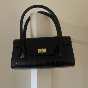 Svart handväska med krokodilmönster - Elegant svart handväska med krokodilmönster och guldfärgad låsdetalj. Väskan har dubbla handtag och en stilren design som passar perfekt för en chic look.