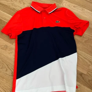Flerfärgad pikétröja från Lacoste - Snygg pikétröja från Lacoste i rött, blått och vitt. Tröjan har en klassisk krage med knappar och det ikoniska Lacoste-märket på bröstet. Perfekt för en sportig look!
