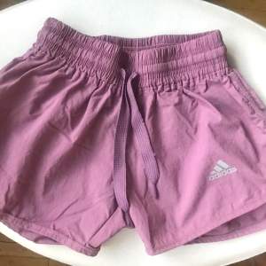 Snygga lila träningsshorts från Adidas med elastisk midja och dragsko för perfekt passform. Perfekta för sommarens alla aktiviteter. Adidas-logga i vitt på benet ger en sportig touch.