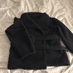 Svart ulljacka med bälte - Svart ulljacka med bälte i midjan. Jackan har en oversized passform och långa ärmar. Använd ett fåtal gånger