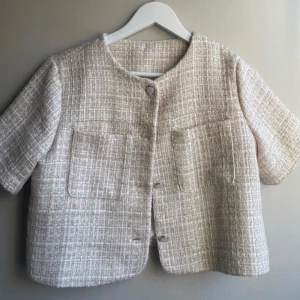 Beige kavaj med kort ärm - Säljer en stilren beige kavaj med korta ärmar och knappar framtill. Kavajen har en elegant, strukturerad yta och är perfekt för en chic look. Passar bra till både jeans och kjol.