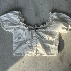Vit blus från Gina Tricot - Säljer en söt vit topp från Gina Tricot med knappar framtill och volangdetaljer vid ärmslut och hals. Blusen har korta puffärmar och en elastisk halslinning som ger en romantisk look. Perfekt för varma dagar!