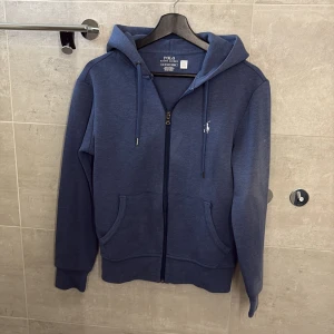 Blå hoodie från Ralph Lauren - Säljer en snygg blå hoodie från Ralph Lauren med dragkedja och huva. Tröjan har långa ärmar och en liten logga på bröstet. Perfekt för en avslappnad stil. Inte det ”vanliga” hoodie materialet utan det som är lite ”exklusivare” köpte den helt ny för 2099kr