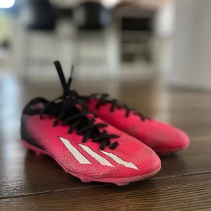 Rosa och svarta fotbollsskor från Adidas - Snygga fotbollsskor i rosa och svart med vita detaljer från Adidas. Skorna har snörning och en sportig design som passar perfekt för fotbollsplanen.