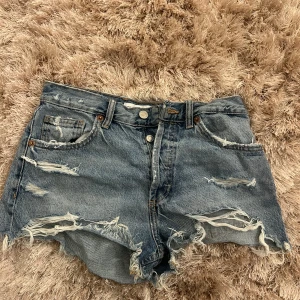 Blå jeansshorts med slitningar - Säljer ett par coola blå jeansshorts med slitna detaljer och hög midja. Shortsen har en klassisk femficksdesign och knappar framtill. Perfekta för en avslappnad stil.