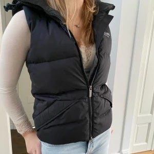 Svart dunväst från Kingsland - Snygg svart dunväst från Kingsland med dragkedja och huva. Perfekt för kyligare dagar och ger en sportig look. Västen har en normal passform och är idealisk för ridning eller andra utomhusaktiviteter.