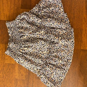 Blommig kjol från H&M - Säljer en söt blommig kjol från H&M i storlek S.Kjolen har ett elastiskt midjeband och ett härligt blommönster i lila, svart och orange. Perfekt för en somrig look!
