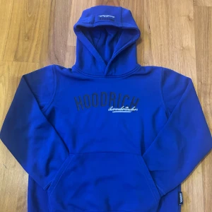 Blå hoodie från Hoodrich - Säljer en snygg blå hoodie från Hoodrich med tryck på bröstet och ryggen. Den har en klassisk känguruficka och en bekväm huva. Perfekt för en avslappnad stil. Passar ungdomar i åldern 12-13 år.