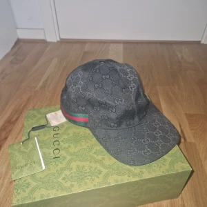 Svart keps från Gucci - Snygg svart keps från Gucci med det ikoniska GG-mönstret. Kepsen har en röd och grön randdetalj på sidan. Perfekt för att ge din outfit en lyxig touch.
