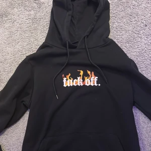 Svart hoodie från FSBN - Säljer en svart hoodie från FSBN med tryck på bröstet som har texten 'fuck off' i vit med eldflammor. Hoodien har en klassisk design med dragsko i huvan och en bekväm passform.