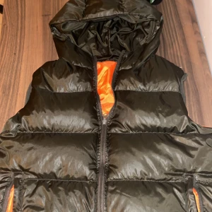 Svart dunväst med orange foder - Snygg svart dunväst med en cool orange insida. Västen har en dragkedja framtill och en huva för extra värme. Perfekt för kyliga dagar när du vill hålla stilen.  Pris kan diskuteras 