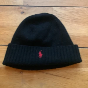 Svart mössa från Polo Ralph Lauren (10-12 år) - Snygg svart mössa från Polo Ralph Lauren med röd broderad logotyp framtill. Tillverkad i mjukt material med ribbad kant för extra komfort. Perfekt för kyliga dagar. Passar 10-12 åringar, för fler frågor/bilder/pris kontakta mig :)