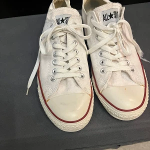 Vita sneakers från Converse - Klassiska vita Converse All Star sneakers med snörning. Skorna har en låg profil och är dekorerade med röda och blå ränder runt sulan. Perfekta för en avslappnad stil. Har spruckit lite på högerskon (se bild)