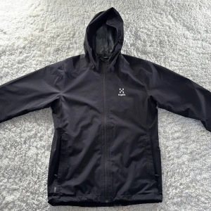 Svart vindjacka från Haglöfs - Säljer en stilren svart vind/regn jacka från Haglöfs. Jackan har en praktisk huva och dragkedja framtill. Perfekt för blåsiga eller regniga vår/höst dagar och har en enkel design med Haglöfs logga på bröstet. Är använd en del men är i väldigt fint skick.