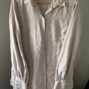 Beige långärmad skjorta - Snygg beige skjorta med långa ärmar och klassisk krage. Skjortan har knappar framtill och vid ärmsluten, vilket ger en stilren look. Perfekt för en avslappnad men ändå uppklädd stil.