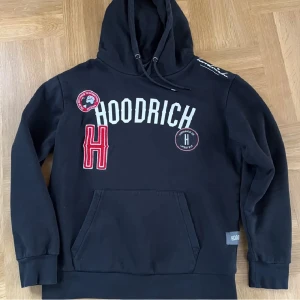 Svart hoodie från Hoodrich - Säljer en svart hoodie från Hoodrich med tryck och broderade detaljer i vitt och rött. Den har en stor ficka framtill och justerbar huva. Perfekt för en casual look.