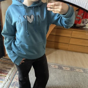 Blå hoodie med hjärtdetalj - Säljer en blå hoodie med en unik hjärtdetalj på bröstet. Tröjan har en känguruficka och justerbar huva med dragsko. Perfekt för en avslappnad stil.