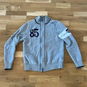 Grå stickad tröja från Tommy Hilfiger - Säljer en snygg grå stickad tröja från Tommy Hilfiger med dragkedja och broderad siffra '85' på bröstet. Tröjan har långa ärmar och ribbade muddar. Perfekt för en stilren och avslappnad look.