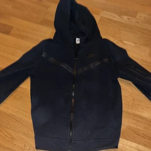 Svart nike tech - Säljer en svart huvtröja från Nike med dragkedja och huva. Tröjan har en diskret logga på bröstet och en praktisk ficka med dragkedja på ärmen. Den är i barn storlek L men passar som XS