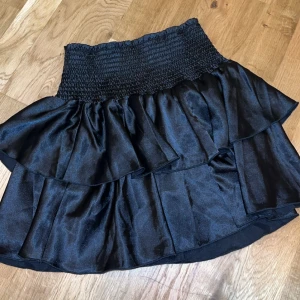 Svart volangkjol - Snygg svart kortkjol med volanger och elastisk midja. Kjolen har en glansig finish. I storlek S