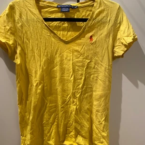 Gul t-shirt från Ralph Lauren Sport - Säljer en gul t-shirt från Ralph Lauren Sport i 100% bomull. Den har en v-ringning och korta ärmar med en broderad logotyp på bröstet. Perfekt för en avslappnad stil.