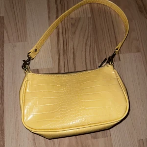 Gul axelväska från Gina Tricot - Snygg gul axelväska i syntetmaterial med krokodilpräglat mönster. Väskan har en praktisk dragkedja upptill och en justerbar axelrem. Perfekt för att liva upp din outfit med en pop av färg!