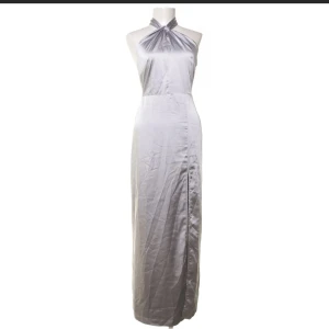 Silverfärgad långklänning från BikBok med slits - Elegant silverfärgad långklänning från BikBok designad av Lovisa Barkman. Klänningen har en öppen rygg och en halterneck-design med en fin rosett i nacken. Perfekt till balen med en snygg slits på sidan. Klänningen är i nyskick och har aldrig använts.