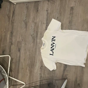 Vit t-shirt från Lanvin - Säljer en stilren vit t-shirt från Lanvin med svart text på bröstet. Perfekt för en minimalistisk look. T-shirten har en klassisk rund hals och korta ärmar.