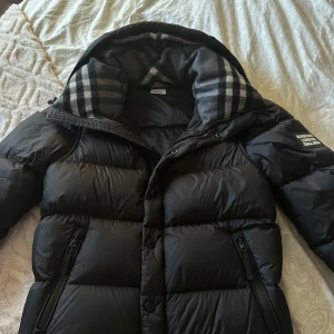 Svart pufferjacka med avtagbara ärmar - Snygg svart pufferjacka från Burberry med avtagbara ärmar och klassiskt rutigt foder i huvan. Jackan har dragkedja och knappar framtill samt praktiska fickor med dragkedja. Perfekt för kyliga dagar när du vill ha flexibilitet i din stil.