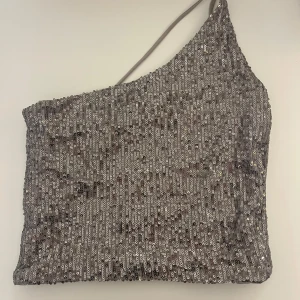 Glittrig oneshoulder topp - Snygg glittrig topp med oneshoulder-design och tunna axelband. Perfekt för en festlig. Toppen är täckt med paljetter som ger en lyxig känsla. Omsydd från storlek M till S/XS. Använd en gång.