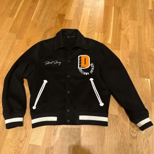 Svart jacka från Zara - Snygg svart jacka från Zara med vita detaljer och broderad text. Jackan har långa ärmar och knappar framtill. Perfekt för en stilren look.