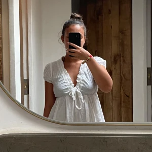Vit klänning med knytning - Charmig vit klänning med knytning framtill och korta ärmar. Perfekt för en avslappnad sommarlook. Den har en v-ringad design och är tillverkad i ett lätt material.