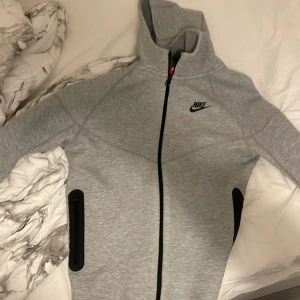 Grå huvtröja från Nike - Säljer en grå huvtröja från Nike med dragkedja och svarta detaljer. Tröjan har en justerbar huva och långa ärmar, perfekt för en sportig look. Nike-loggan är tryckt på bröstet. Passar bra för kyligare dagar.