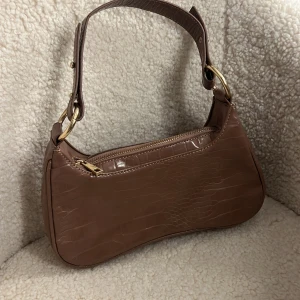 Brun axelväska från NLY Accessories - Snygg brun axelväska i syntetmaterial med krokodilpräglat mönster. Väskan har en justerbar axelrem och en dragkedja upptill för säker förvaring. Perfekt för att ge en stilren touch till din outfit.