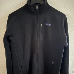 Svart fleecejacka från Patagonia - Säljer en svart fleecejacka från Patagonia med dragkedja framtill. Jackan har en hög krage och långa ärmar, perfekt för kyliga dagar. Den är stilren och enkel att matcha med andra plagg.