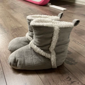 Gråa tofflor med fluffig insida - Mysiga gråa tofflor från Sweet Dreams med en fluffig vit insida. Perfekta för att hålla fötterna varma under kalla dagar. De har en mjuk och bekväm design med en liten flik baktill för enkel påtagning.