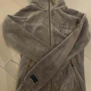 Grå fleecejacka från Fjällräven - Mysig grå fleecejacka från jacson med dragkedja och huva. Perfekt för kyliga dagar. Jackan har långa ärmar och praktiska fickor med dragkedja.