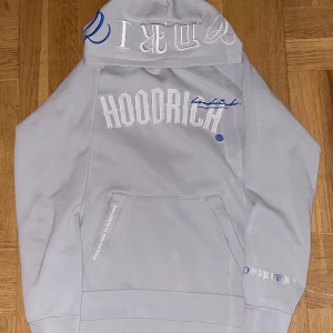  Hoodrich - Snygg grå hoodie från Hoodrich med vit och blå textdetalj på framsidan. Den har en stor ficka framtill och text på ryggen. Perfekt för en avslappnad stil.