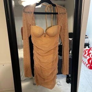 Beige klänning med pärldetaljer oh polly - Elegant beige klänning med vadderade kupor och pärldetaljer på de långärmade ärmarna. Klänningen har en rynkad design och en dekorativ pärla framtill. Perfekt för en festlig kväll.