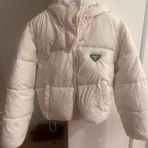 Vit pufferjacka från Prada - Säljer en snygg vit pufferjacka från Prada med huva och dragkedja. Jackan har en klassisk design med tryckknappar och två fickor framtill. Perfekt för kyliga dagar.