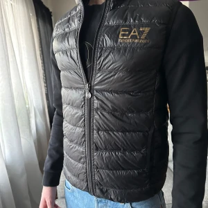 Svart dunväst från Emporio Armani - Säljer en snygg svartglansig dunväst från Emporio Armani med dragkedja och logga i guld på bröstet. Perfekt för lager-på-lager under kyliga dagar. Använd ett fåtal gånger och har en bra passform. Hör av dig för fler bilder och specifika mått 💫 