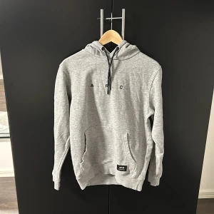 Grå hoodie från !Solid - Säljer en stilren grå hoodie från !Solid med tryck 'A.R.C' på bröstet. Tröjan har en klassisk känguruficka och justerbar huva med snören. Perfekt för en avslappnad stil.
