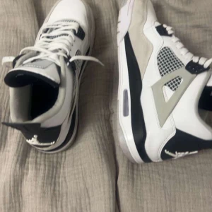 Nike Air Jordans i vitt och svart ( air Jordan 4 ) - Snygga Nike Air Jordans i vitt och svart med grå detaljer. Skorna har klassisk snörning och en ikonisk design med synlig luftdämpning i sulan. Perfekta för dig som vill ha stil och komfort i ett.