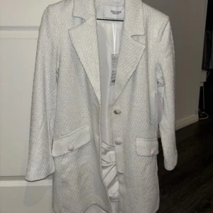 Vit kavaj  - Elegant vit kavaj med texturerat tyg och klassisk krage. Kavajen har långa ärmar och knappar framtill samt fickor med lock. Perfekt för en stilren look.